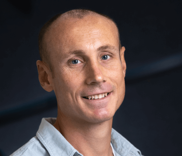 Photo of white man in blue shirt smiling against a dark blue background