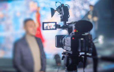 photo of a video camera in foreground and man standing in background wearing a blazer. He is out of focus.